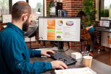 Sakallı genç erkek analist ofis masasındaki bilgisayar ekranında mali tablolara bakıyor. Tuğla duvar odasında oturan profesyonel işadamı, başlangıç şirketinin pazar analizi için masaüstü bilgisayarını kullanıyor.