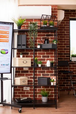 İş analitiğini özetleyen dijital ekran görüntülemesi olarak bitki süslemeleri ve kutularla düzenlenmiş ofis dolabı. Tuğla duvara yerleştirilmiş iyi örgütlenmiş raf içeren modern çalışma alanı.
