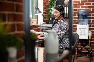 Organize bir girişim ofisinde, masaüstü bilgisayarında kulaklık kullanan kadın girişimci. Bilgisayarında dinleme cihazı olan Asyalı kadın danışman ve müşterilerle konuşuyor..