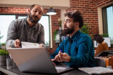 Pano sahibi erkek danışman, masa başında dizüstü bilgisayarla oturan iş arkadaşıyla pazarlama stratejisini analiz ediyor. Kafkasyalı işadamları, modern çalışma alanındaki temel şirket hedeflerine ulaşmak için işbirliği yapıyorlar..