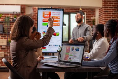 El kaldırılmış bir iş kadını dizüstü bilgisayarında oturmuş veri tablolarını gösteriyor ve iş arkadaşlarıyla strateji planı toplantısında sorular soruyor. Beyaz bir bayan kolunu kaldırıp, fikirlerini takım arkadaşlarıyla paylaşıyor..