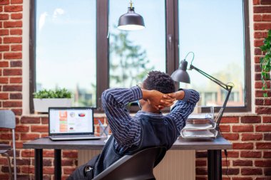 Siyahi girişimci, görevlerini tamamladıktan sonra kendini başarılı hissederek modern başlangıç ofisinde sırtını uzatıyor. Yüz maskeli bir erkek analist, sandalyesine yaslanıyor ve işte verimli bir gün geçirdiğini düşünüyor..