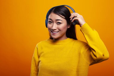 Kulağının yanında kulaklık tutan, mutluluktan gülümseyen, turuncu arka planda duran güzel Asyalı bir kadın. Genç bir bayan kameraya bakıyor ve kablosuz kulaklığına dokunuyor..