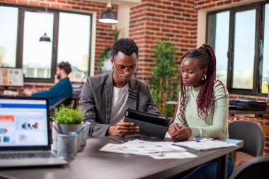 İş adamı, tabletteki pazarlama stratejisini tuğla duvar çalışma alanındaki kadın çalışana açıklıyor. Afrika kökenli Amerikalı meslektaşlar masada belgelerle oturuyor ve ofiste ciddi bir konuşma yapıyorlar..