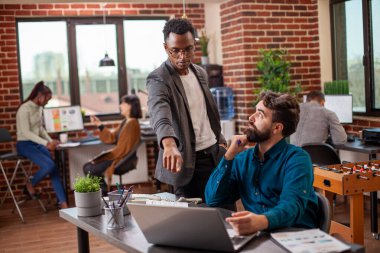 Erkek analist, beyaz iş arkadaşına pazar analizi güncellemesini açıklayan dizüstü bilgisayarını işaret ediyor. Sakallı adam oturur ve siyahi yöneticinin sunumunda yaptığı finansal hataları dikkatle dinler..