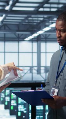 Veri merkezi yöneticilerinden oluşan dikey video ekibi evrak dosyalarını okuyor, sistem tanılarını gözden geçiriyor. Sunucu odası çalışanları bakım programı belgelerini ve siber güvenlik protokollerini inceliyor.