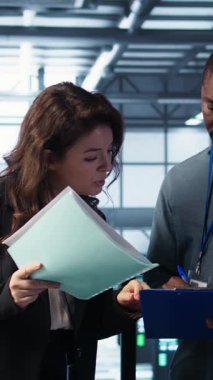 Dikey video veri merkezi yöneticileri evrak dosyalarını okuyor, sistem tanılarını gözden geçiriyorlar. Sunucu çiftliği meslektaşları bakım takvimlerine, belgelere ve siber güvenlik protokollerine bakıyor.