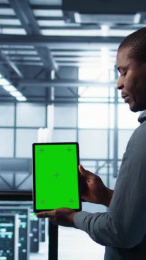 Dikey video sunucu odası veri bilimcileri sahte tablet kullanıyor, hataları önlemek için güvenlik denetimleri yapıyor. Veri merkezinde iş arkadaşı olan Afrikalı Amerikalı bir adam yeşil ekran cihazına bakıyor, kamera B