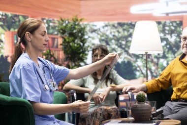 Röntgen tarama sonuçlarını ve teşhisini sunan sağlık görevlisi yeni sağlık tedavisini tartışıyor. Hemşire, tekerlekli sandalyedeki yaşlı kadına ve kocaya radyografiyi gösteriyor..