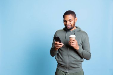 Confident optimistic man browsing his smartphone and serving coffee, feeling happy after receiving good news and socializing with his friends via text messages. Modern guy with a refreshment. clipart