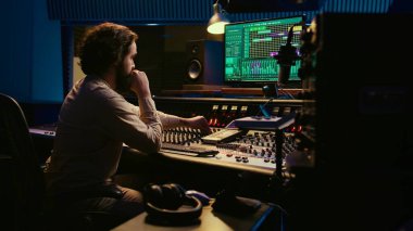 Müzik yapımcısı kabloyu amfi ya da ses kompresörünün içinde tamir ediyor, kontrol odası teknik ekipmanlarını kayıt seansı için hazırlıyor. Ses mühendisi şarkılar üzerinde çalışıyor. Kamera B.