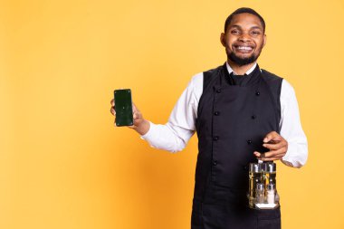 Caterer with apron and cruet set shows green screen on smartphone, working in catering customer focused industry. Professional butler in uniform holds phone with mockup display. clipart