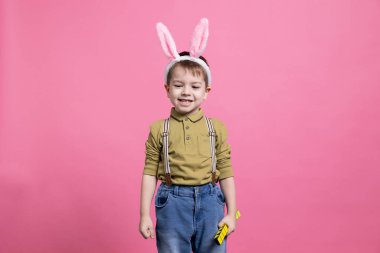 Paskalya bayramını kutlamak için tavşan kulağı takarken kameranın önünde gülümseyen masum bir çocuk. Pembe arka planda poz veren neşeli küçük çocuk, şirin renkli oyuncak..