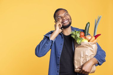 Genç erkek manken ailesiyle telefonda konuşuyor ve market poşeti taşıyor, taze organik ürün alışverişi yapıyor ve akıllı telefondan arkadaşlarıyla sohbet ediyor. Toptan ürünlerle gülümseyen adam..