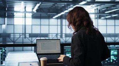 Veri merkezinde dizüstü bilgisayar kullanan programcı, ağ protokollerini ayarlayan, kahve içen. Sunucu çiftliğindeki kadın defter üzerinde kodlama yapıyor, içeceklerden hoşlanıyor, gelişmiş güvenlik önlemleri uyguluyor.