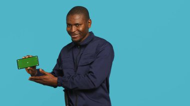 Portrait of upbeat security staff member showing green screen smartphone. Cheerful patrolling agent maintains secure environment, showcasing mockup phone, studio background, camera A clipart