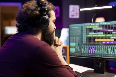 Müzik mühendisi, ev stüdyosuna yeni ses efektleri ekledikten sonra kulaklıkla ses kaydını dinliyor. Yapımcı, müzik hacmini ve stereo aracını geliştirmek için pc üzerinde daw yazılım arayüzü kullanıyor.