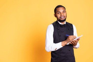 Professional butler writing the order of the clients on notepad, takes notes to serve five star restaurant customers. African american waiter wears apron, service oriented industry. clipart