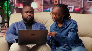 Afro-Amerikan çalışanlar oturma odasında dizüstü bilgisayar kullanarak sunum hazırlıyor, çizelgeler yerleştiriyor ve düzenleri ayarlıyorlar. Genç çift şirket projesini bitiriyor ve özetler yazıyor. Kamera B.