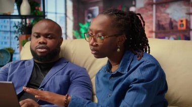 Afro-Amerikan çalışanlar oturma odasında dizüstü bilgisayar kullanarak sunum hazırlıyor, çizelgeler yerleştiriyor ve düzenleri ayarlıyorlar. Genç çift şirket projesini bitiriyor ve özetler yazıyor. Kamera A.