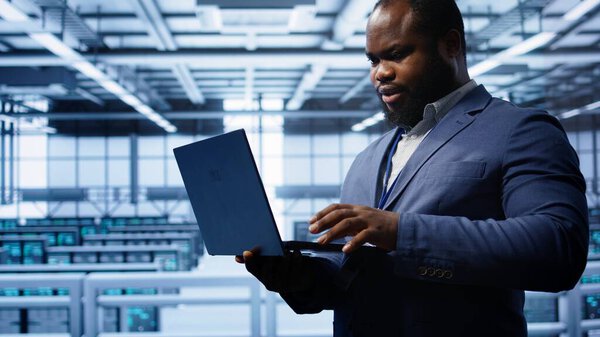 Computer scientist in server hub checking recovery plan on laptop, monitoring hardware energy consumption. IT specialist uses notebook in data center facility, ensuring rigs are functioning, camera B