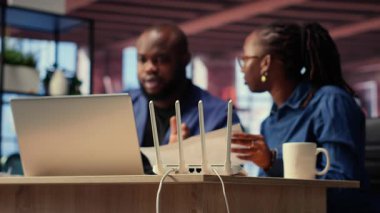 Siyah sinir bozucu erkek ve kadın kablosuz yönlendiricilerini yeniden başlatıp ağ ayarlarını kontrol ediyorlar, bir internet kesintisini çözme çabalarına kızıyorlar ve görevlerini aksatıyorlar. Kamera B.