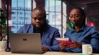 Defterdeki hesap çizelgelerini tamamlayan, hücrelere sayısal verileri giren, toplamları hesaplayan bir kadın. Afrikalı Amerikalı çift, bir muhasebe görevi için internet bilgilerini inceliyor. Kamera B.