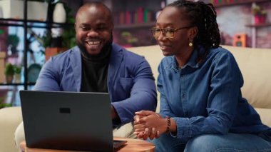 Siyahi genç bir çift evlerinde video bağlantısı kuruyor, kanepede oturuyor ve arkadaşlarıyla konuşuyor. Kadın ve erkek video konferansında el sallıyor, yaşam tarzı. Kamera B.