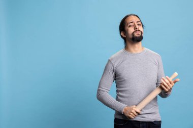 Stüdyoda birini merdaneyle dövmekle tehdit eden agresif bir genç adam kamerada dövüşmeye hazırlanıyor. Sinirli yetişkin, mavi arka planda şiddete başvuruyor..