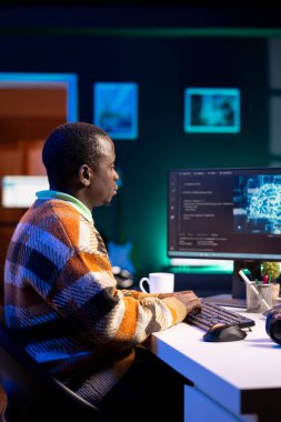 Afro-amerikan bilişim uzmanı makine öğrenimi için yapay zeka sistemini yönetiyor, terminal penceresinde kodlama yazılımı var. Gelecekteki arayüz programı veritabanı için siber güvenlik ve ağ oluşturmaya odaklanıyor.