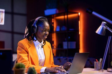 Uzaktan müzik dinleyen, masadaki bilgisayardaki verileri analiz eden, analitik raporları derleyen bir çalışan. Ofisteki kadın kulaklık takıyor, defter cihazı kullanıyor, bilgi yazıyor.