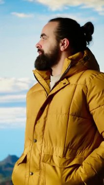 Sıcak kıyafetler giymiş dikey Video Yürüyüşçüsü zirveye tırmandıktan sonra bir dağın tepesinde duruyor, engebeli tepelerin ve engin gökyüzünün nefes kesici manzarasına bakıyor. Manzaralı macera. Kamera.