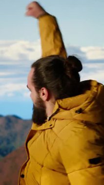 Dikey Video Triumphant yürüyüşçüsü kollarını zaferle kaldırarak dağın zirvesinde duruyor ve başka bir dağa tırmanmanın başarısını ve kişisel başarısını kutluyor. Nefes kesici panoramik manzara