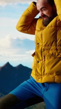 Dikey Videolu Erkek doğa aşığı dağın tepesinde soğuk rüzgarla boğuşuyor, kayalık tepelere tırmanıyor ve üşüyor. Genç yürüyüşçü maceracı manzaranın tadını çıkarıyor.