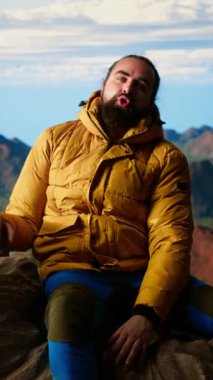 Dikey Video Erkek dağ yürüyüşçüsü muzaffer bir tırmanışı kutluyor ve kolları heyecan içinde kayaların üzerinde otururken dinleniyor. Manzaralı bir arka planda panoramik manzara ve temiz hava