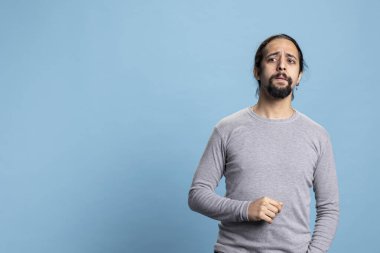 Ciddi bir insan aynı anda hem meraklı hem de şüpheci davranıyor, günlük kıyafetler giyiyor ve mavi arka plana karşı poz veriyor. Uzun kıvırcık saçlı ve sakallı genç yetişkin başka bir şeye odaklanıyor..
