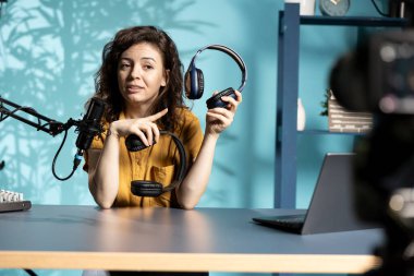 Ses mühendisi, internet kanalı için video kaydediyor. Parlak ışıklandırılmış ev stüdyosunda seyircilere oyun kulaklığı sunuyor. İzleyicilere müzik dinleme aygıtı gösteren kadın