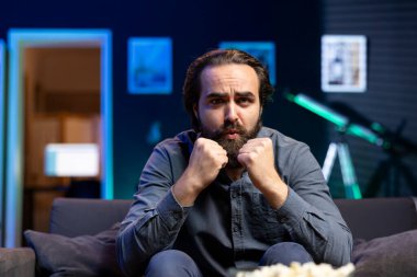 Televizyonda dizi izleyen, yumruklarını sıkan, karakterleri destekleyen, ekrana dikkatle bakan bir adam. Adam dairede koltukta oturup el sıkışarak mutlu sonla bitmesini umuyor.