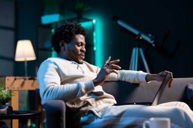 Mutlu adam sıcak oturma odasında dizüstü bilgisayardaki komik internet videolarını izleyerek rahatlıyor. Neşeli insan, neon ışıklı dairede boş vakitlerin tadını çıkarıyor ve online videolara bakıyor.