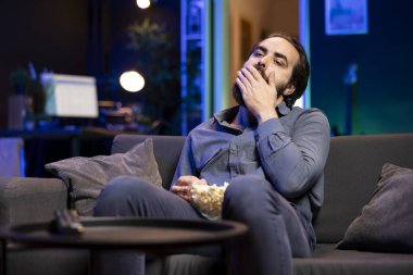 Evde kanepede gevşeyen sıkkın bir adam, evde televizyon izlerken kaseden çıkan patlamış mısırı ağzına tıkıyor. RGB 'nin aydınlattığı oturma odasında abur cubur yiyen tembel biri.