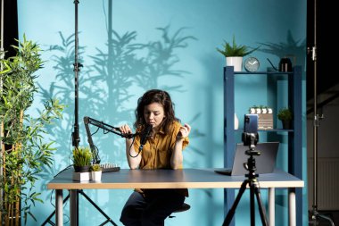 Stüdyoda akıllı telefonuyla video bloğu çeken bir kadın mikrofon ekipmanına konuşuyor. Çevrimiçi kanalda trend başlıkları hakkında abonelere konuşan nüfuzlu kişi, cep telefonuyla kayıt yapıyor