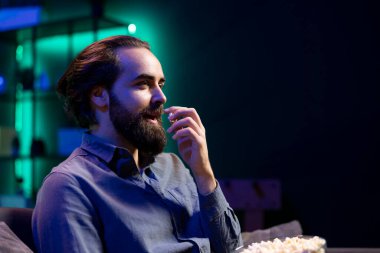 Evde dinlenme saatinde televizyonda kablolu yayın izlerken patlamış mısırın keyfini çıkaran mutlu bir adam. Oturma odasındaki koltukta oturan neşeli izleyiciler atıştırmalık yemek ve televizyon dizilerine bakmak için sabırsızlanıyorlar.