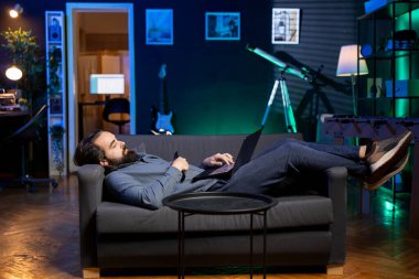 Ev sahibi evde yatıp televizyon izlemekten ve dizüstü bilgisayarda yazmaktan sıkılmış. Adam kanepede uzanmış, dizüstü bilgisayarla akılsızca eğleniyor.