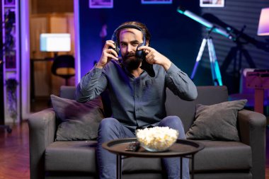 Neon ışıklı dairede video oyunları oynamak için kulaklık takan gülümseyen adam, elinde kontrol cihazı tutuyor. Oyuna konsol sistemi ve kulaklık kullanarak katılan oyuncu