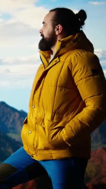 Sıcak kıyafetler giymiş dikey Video Yürüyüşçüsü zirveye tırmandıktan sonra bir dağın tepesinde duruyor, engebeli tepelerin ve engin gökyüzünün nefes kesici manzarasına bakıyor. Manzaralı macera. Kamera.