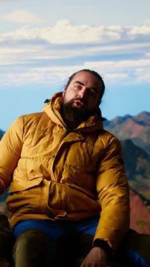 Dikey Videolu Memnuniyet Yürüyüşçüsü dağın zirvesine ulaşıyor ve yorgun, dinleniyor ve kayalık dağ zirvesinde dinleniyor. Genç adam nefes kesici manzaranın tadını çıkarıyor. Kamera B.