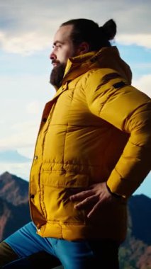 Dikey Video Doğa Aşığı nefes kesen bir dağ sırasının kenarında duruyor, yükselen zirvelerin ve engin gökyüzünün manzaralı arka planına bakıyor. Genç adam macera ve açık hava keşiflerini seviyor.