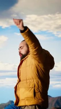 Dikey Video Gezgini yeni zirvelere ulaşıyor ve dağın tepesinde zafer dansı yapıyor, tatmin ve motivasyonu hissediyor. Dağların ve gökyüzünün nefes kesici manzarası macerayı yansıtır.