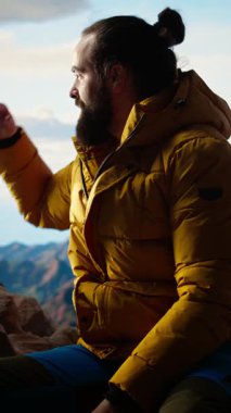 Dikey Video muzaffer yürüyüşçüsü, zirveye tırmandıktan sonra dağda dinleniyor, kişisel başarılarıyla gurur duyuyor. Genç adam kayalarda oturuyor ve huzurlu ortamın tadını çıkarıyor. Kamera.