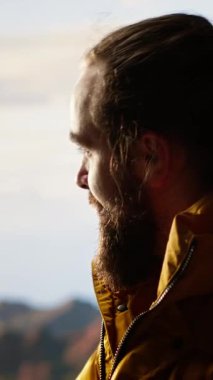 Dikey Video muzaffer yürüyüşçüsü, zirveye tırmandıktan sonra dağda dinleniyor, kişisel başarılarıyla gurur duyuyor. Genç adam kayalarda oturuyor ve huzurlu ortamın tadını çıkarıyor. Kamera.